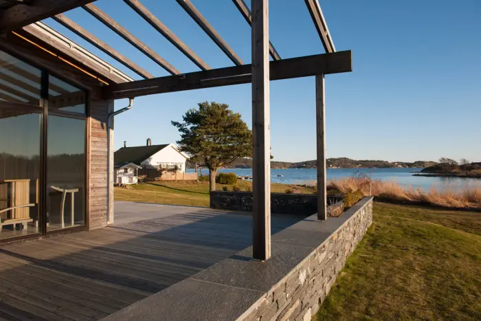 BJØ — terrasse under pergola i tre, med skiferheller og natursteinsmur, mot sjøen og det gamle huset