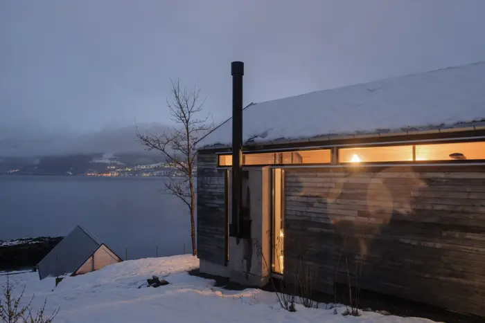 BAL — grånet trefasade med opplyst vindusbånd under taket i skumring, med fjorden bak