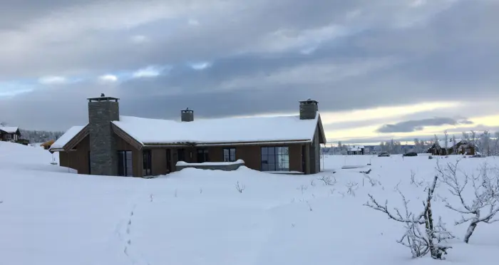 BET — hytte i brun trekledning med to natursteinspiper, i snødekt fjellandskap