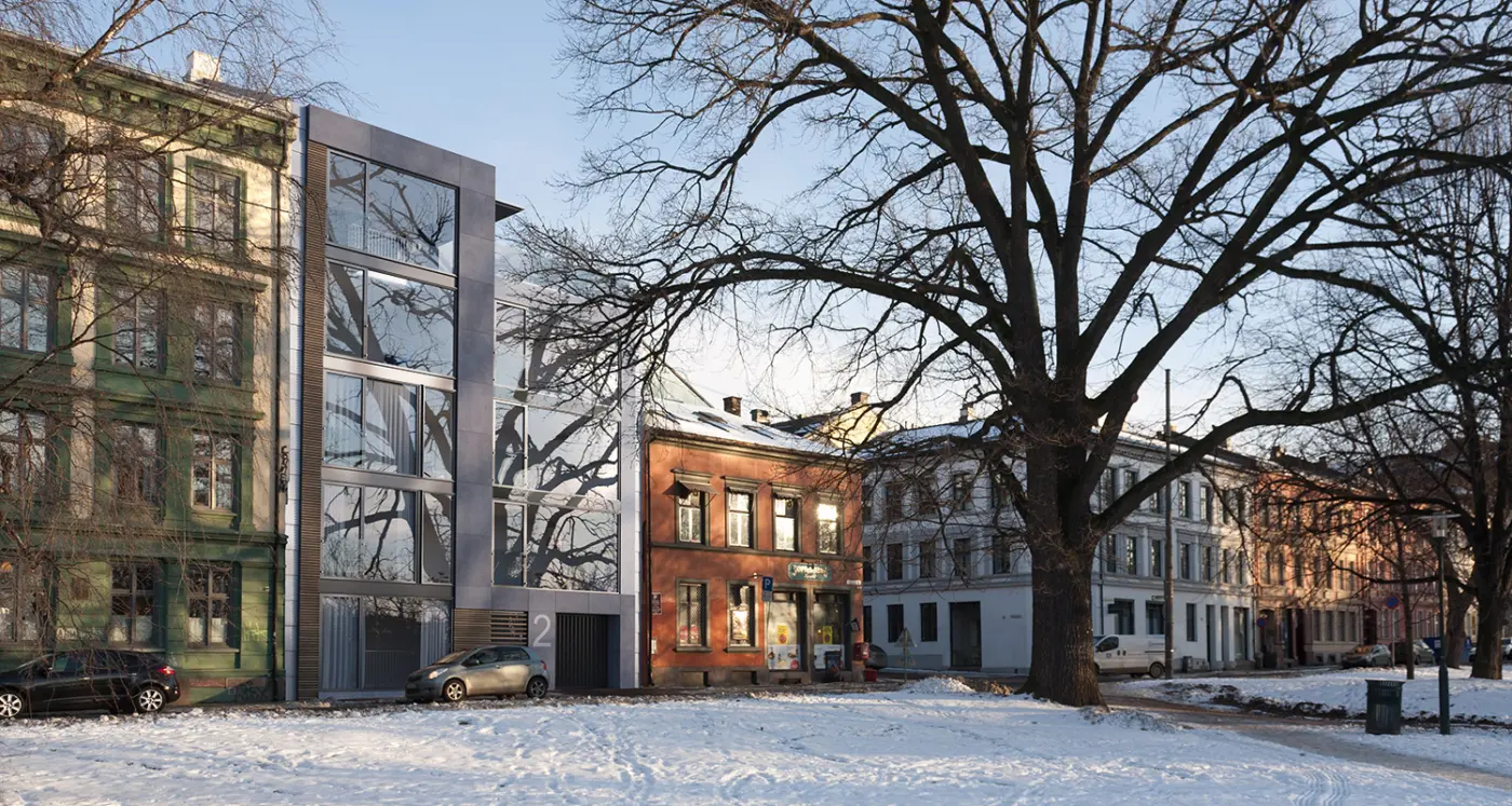HEG42 — smalt nybygg i glass og metall innfelt mellom eldre bygårder, sett fra parken om vinteren