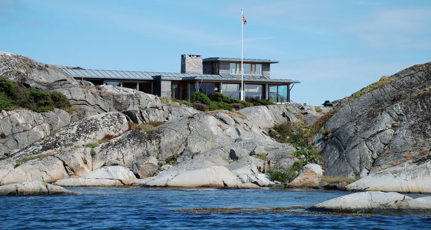 YKH — bolig på svaberg sett fra sjøen, med båndtekket tak, natursteinspipe og flaggstang