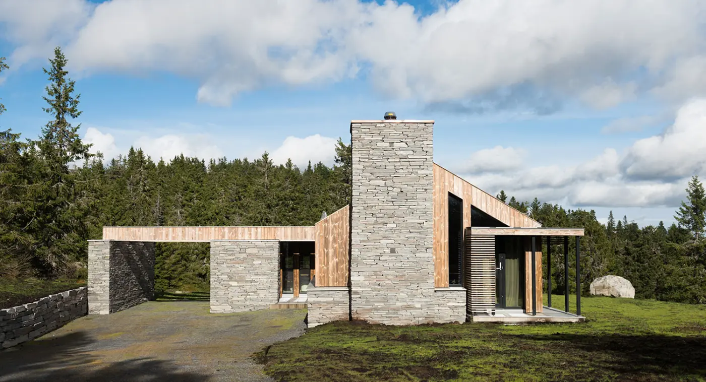SJU — hytte i skifermur og lys trekledning med kraftig natursteinspipe, i granskog