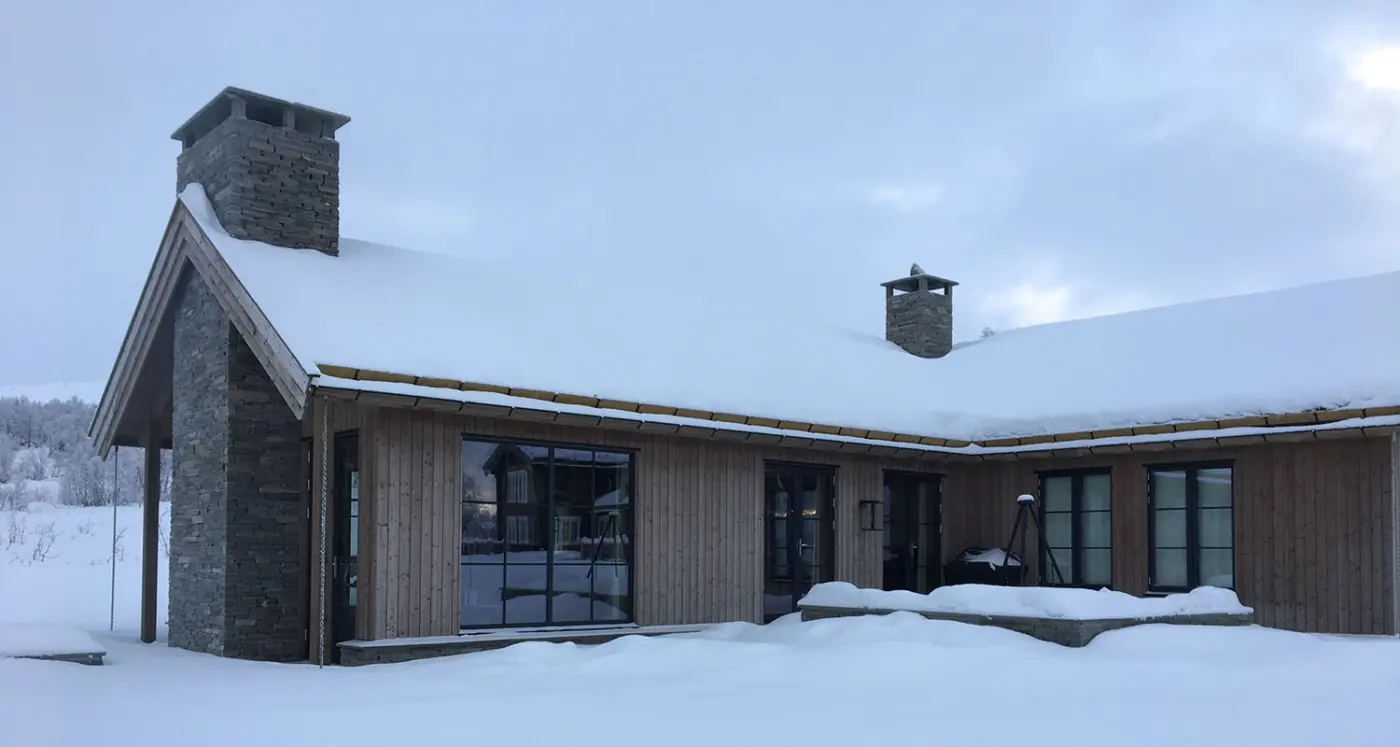 BET — hytte i lys trekledning med saltak og natursteinspipe, snødekt i vinterlandskap