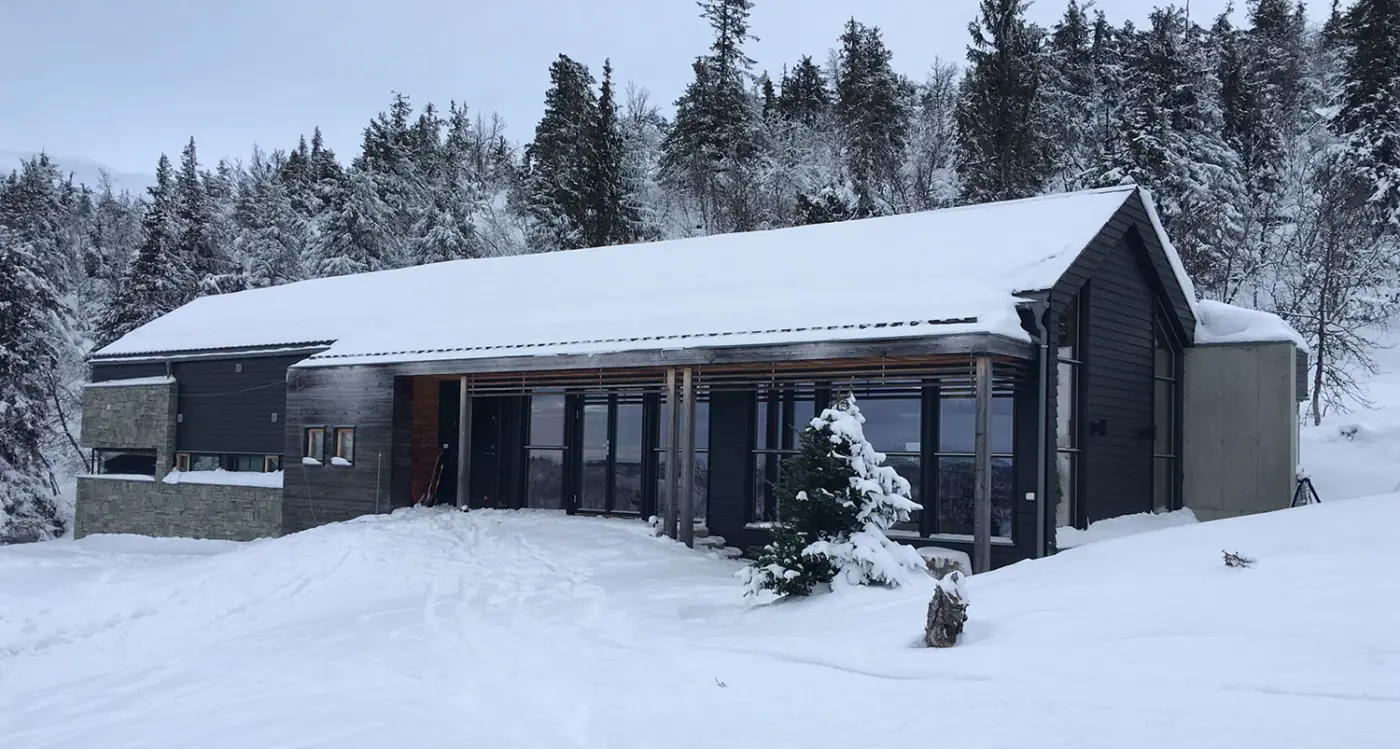 KVF1 — hytte i mørk trekledning med glassfasade bak trestolper, snødekt i granskog