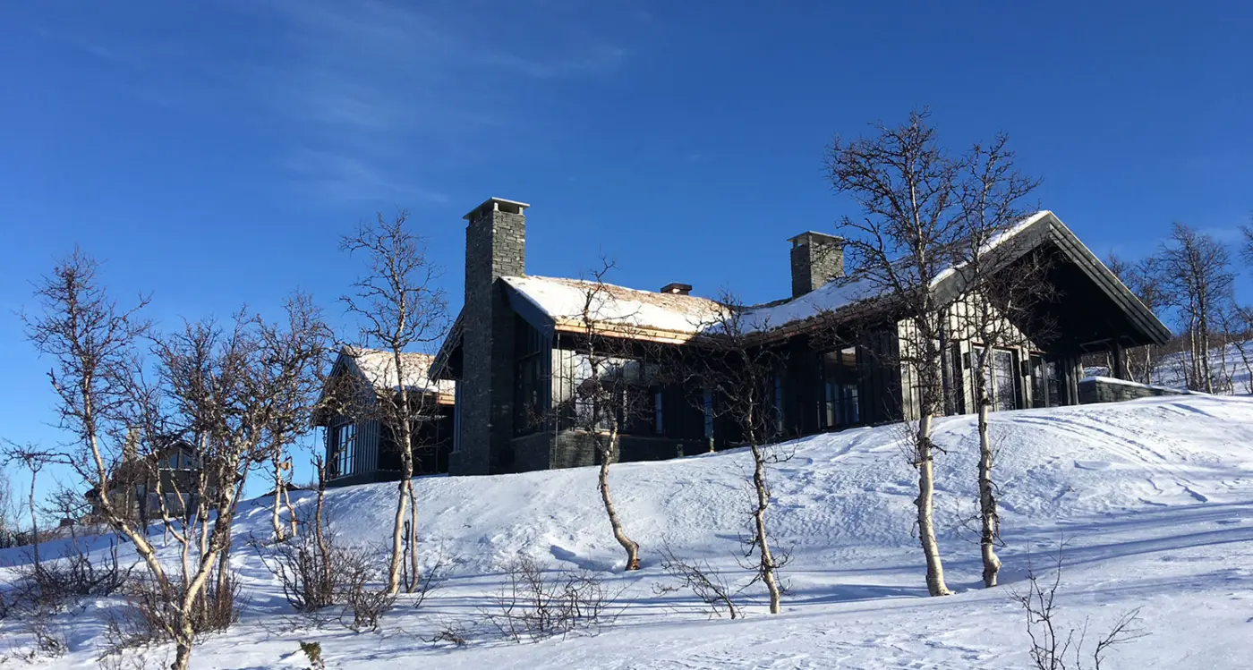 HAV — stor hytte i mørkt tre med torvtak og natursteinspiper, i snødekt bjørkelandskap