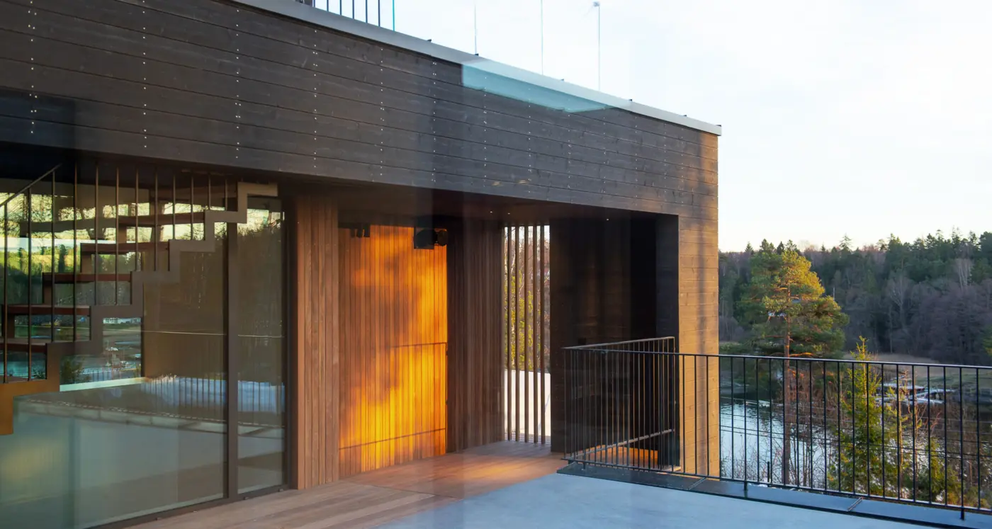 ASK — terrasse i kveldssol med opplyst inngangsparti i tre, med utsikt over vannet og skogen