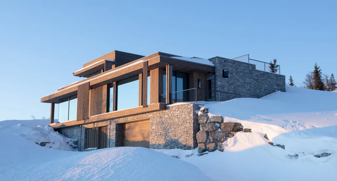 NOR2 — hytte i tre med flate tak over kraftig natursteinsmur og garasje, i kveldssol og dyp snø