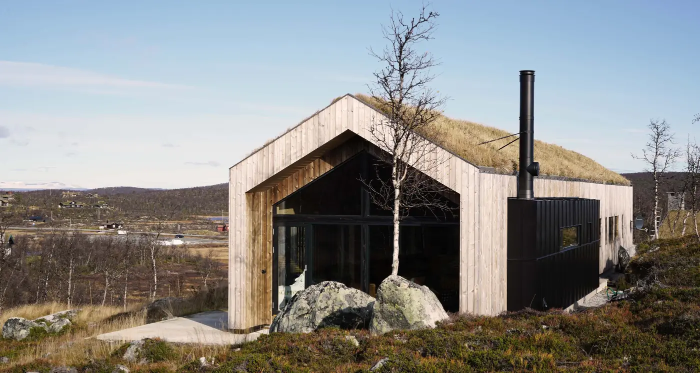 SOL58 — hytte i lys trekledning med torvtak og svart metallkledd fløy, på fjellet med utsikt over dalen