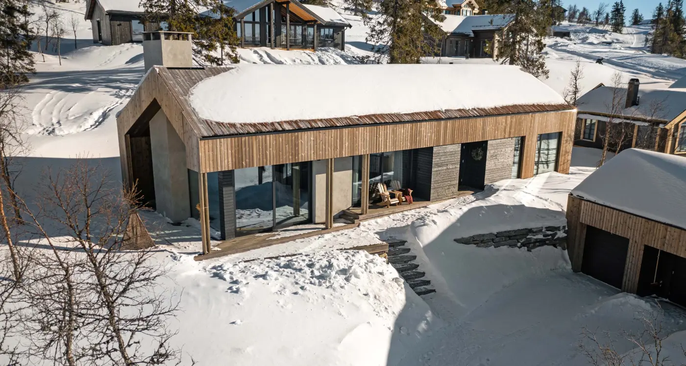 BET125 — hytte i lys trekledning med dypt takutstikk på stolper og terrasse, sett ovenfra i snøen
