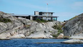 YKH — bolig på svaberg sett fra sjøen, med båndtekket tak, natursteinspipe og flaggstang
