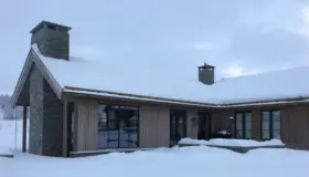 BET — hytte i lys trekledning med saltak og natursteinspipe, snødekt i vinterlandskap