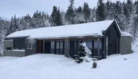 KVF1 — hytte i mørk trekledning med glassfasade bak trestolper, snødekt i granskog
