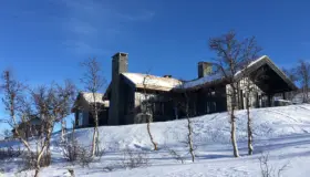 HAV — stor hytte i mørkt tre med torvtak og natursteinspiper, i snødekt bjørkelandskap