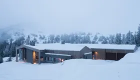 KVF3 — lang lav hytte i tre og naturstein med garasjer, i snøkledd fjellandskap med tåke