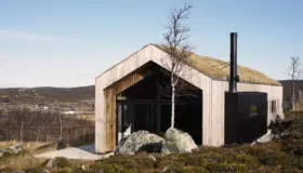 SOL58 — hytte i lys trekledning med torvtak og svart metallkledd fløy, på fjellet med utsikt over dalen