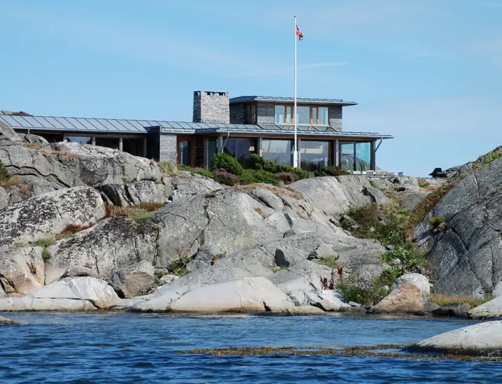 YKH — bolig på svaberg sett fra sjøen, med båndtekket tak, natursteinspipe og flaggstang