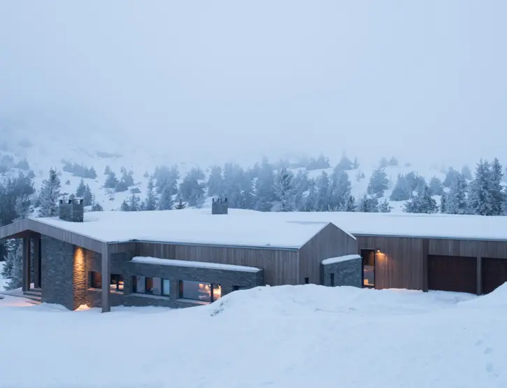 KVF3 — lang lav hytte i tre og naturstein med garasjer, i snøkledd fjellandskap med tåke