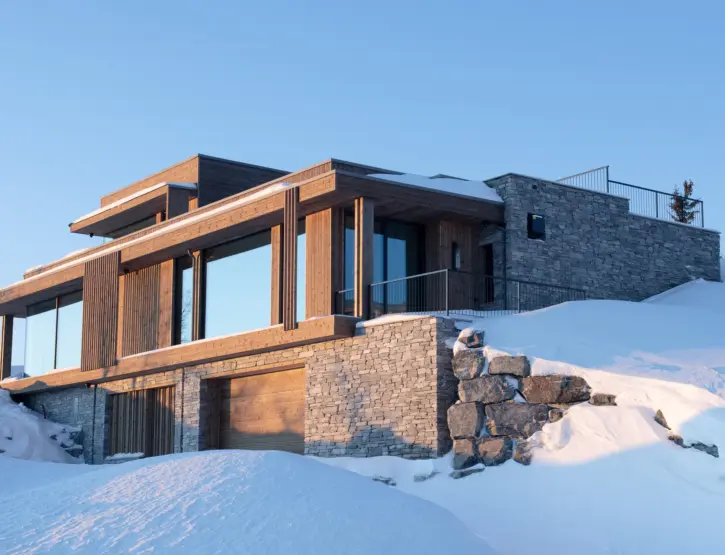 NOR2 — hytte i tre med flate tak over kraftig natursteinsmur og garasje, i kveldssol og dyp snø