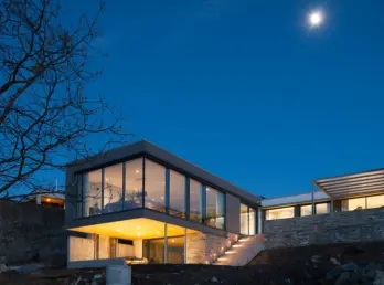 SVV12 — bolig med glassert overetasje utkraget over åpen carport, med natursteinsmur, fotografert i måneskinn