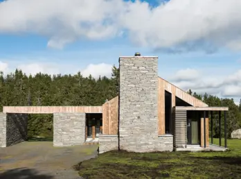 SJU — hytte i skifermur og lys trekledning med kraftig natursteinspipe, i granskog