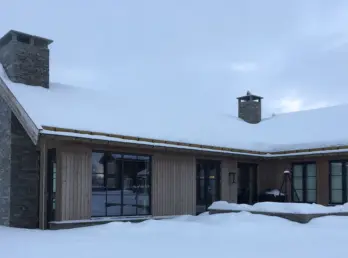 BET — hytte i lys trekledning med saltak og natursteinspipe, snødekt i vinterlandskap