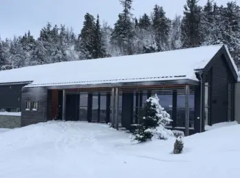 KVF1 — hytte i mørk trekledning med glassfasade bak trestolper, snødekt i granskog