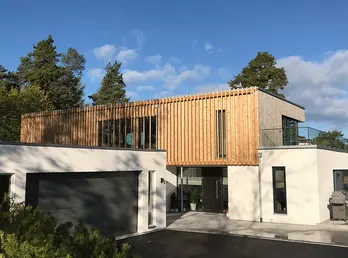 LOV67 — bolig med hvitpusset underetasje og garasje, og overetasje i vertikal trekledning med spilefelt, mot fjorden