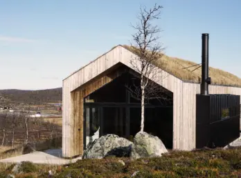 SOL58 — hytte i lys trekledning med torvtak og svart metallkledd fløy, på fjellet med utsikt over dalen