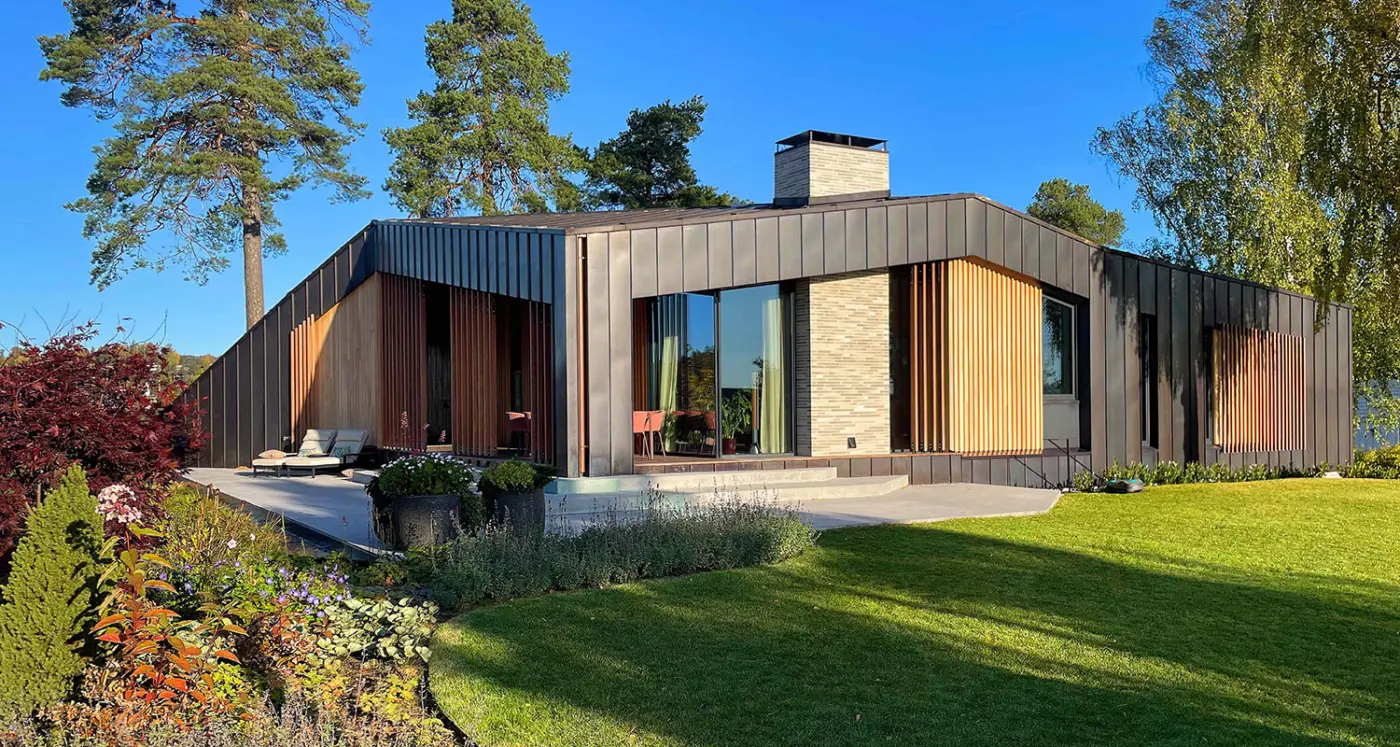 P25 — enetasjes bolig i mørk båndtekket metall med spilefelt i lyst tre og pipe i lys tegl, mot hagen