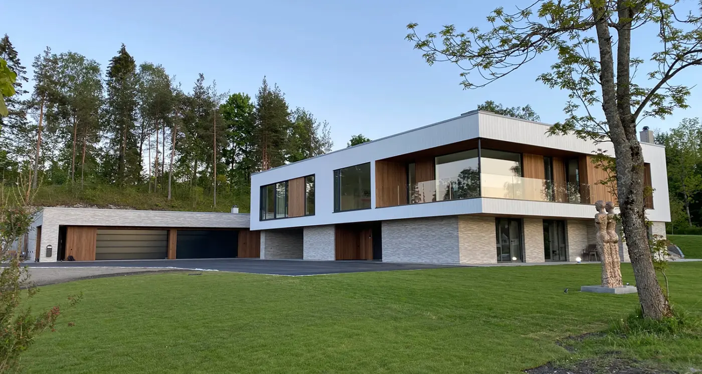 DNV3 — bolig med hvit overetasje over base i lys naturstein, med garasjerekke og skulptur på plenen