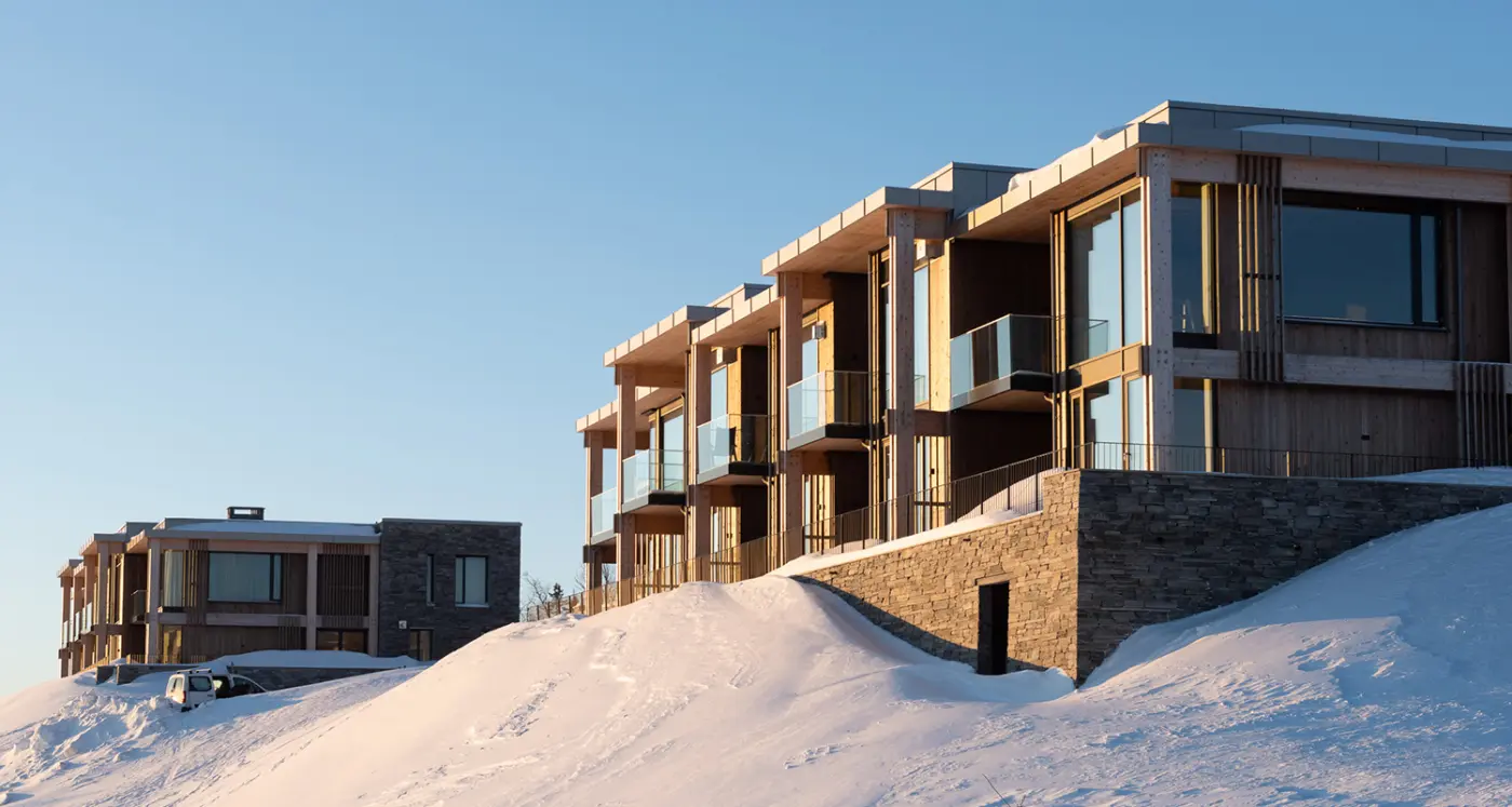 ØNS — leilighetsbygg med trekonstruksjon og glassbalkonger over natursteinsmur, i kveldssol og snø