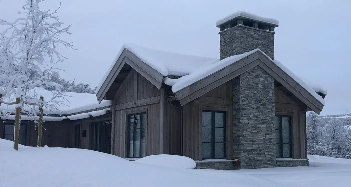 GEI2 — hytte i grånet tre med saltak og natursteinspipe, tungt snødekt