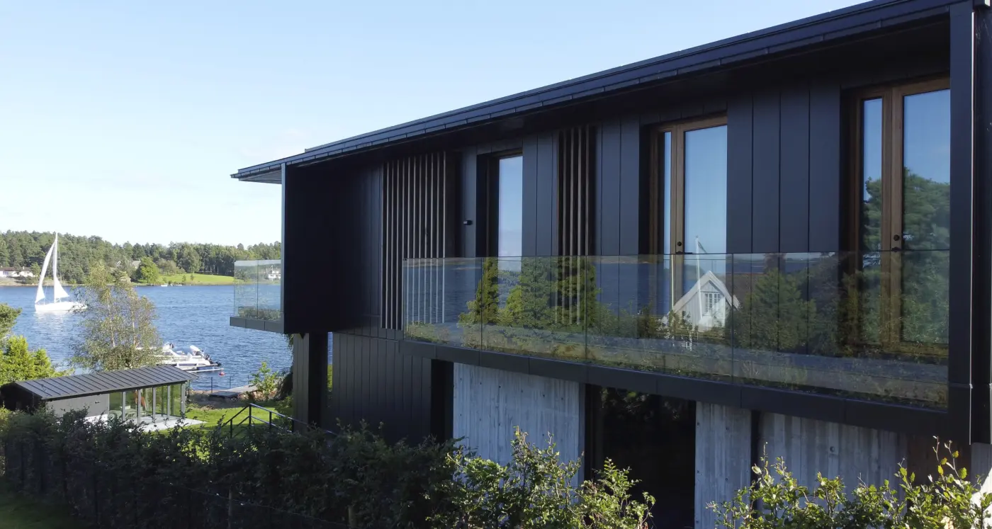 VES — bolig i svart kledning med glassrekkverk over base i forskalingsbetong, med utsikt mot sjøen og naust ved stranden