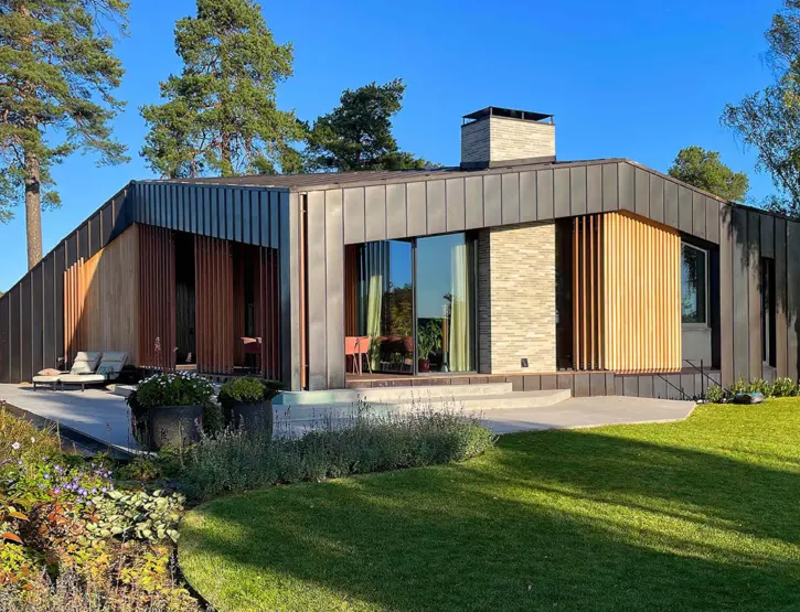 P25 — enetasjes bolig i mørk båndtekket metall med spilefelt i lyst tre og pipe i lys tegl, mot hagen