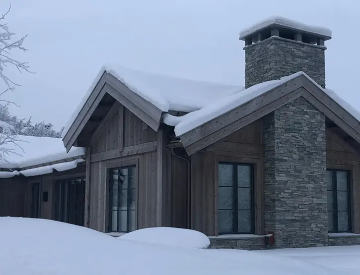 GEI2 — hytte i grånet tre med saltak og natursteinspipe, tungt snødekt