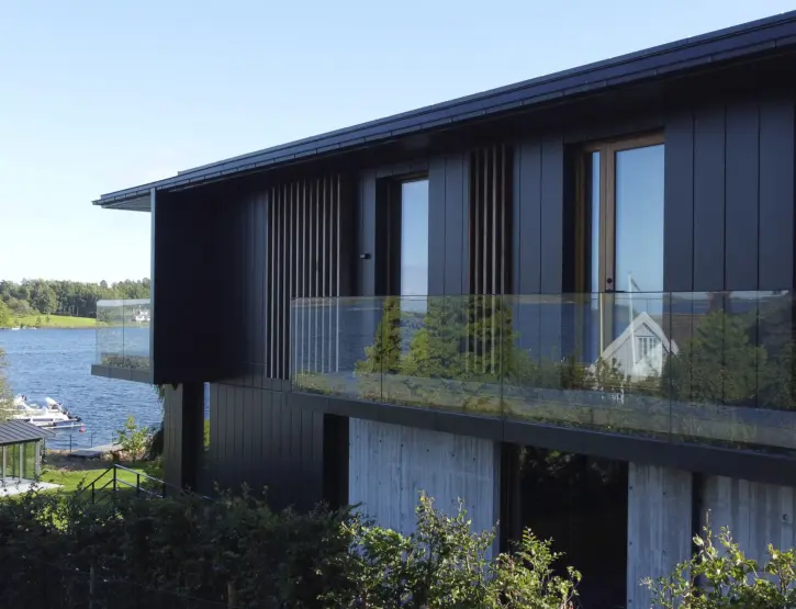 VES — bolig i svart kledning med glassrekkverk over base i forskalingsbetong, med utsikt mot sjøen og naust ved stranden