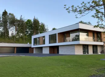 DNV3 — bolig med hvit overetasje over base i lys naturstein, med garasjerekke og skulptur på plenen